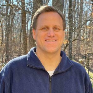 Steve Bensen, rectal cancer survivor, smiles wearing a blue fleece in front of a woodsy background. 